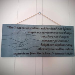 May include: A rectangular, blue-grey wooden sign with a quote from Romans 8:38-39. The sign features a heart held by two hands, with the text in a dark grey font. A twine string is attached for hanging.