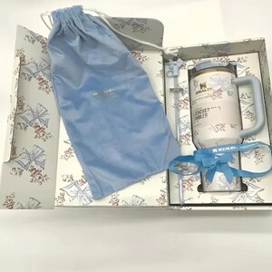 May include: A gift set featuring a white and light blue Stanley Quencher tumbler with a handle, straw, and a blue velvet drawstring bag. The tumbler has the text "ENCHER TUMBLER" and "STANLEY" printed on it. The gift box has a floral pattern.