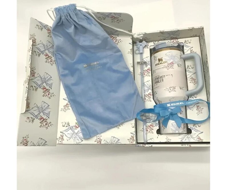 May include: A gift set featuring a white and light blue Stanley Quencher tumbler with a handle, straw, and a blue velvet drawstring bag. The tumbler has the text "ENCHER TUMBLER" and "STANLEY" printed on it. The gift box has a floral pattern.