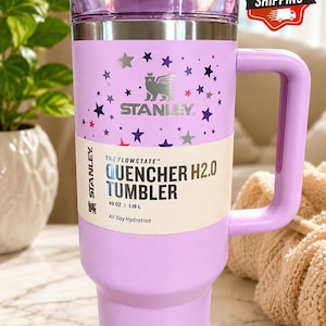 May include: A lavender Stanley Quencher H2.0 tumbler with a handle, lid, and star pattern. The tumbler holds 40 oz (1.18 L) and features the Stanley logo. The text "All Day Hydration" is printed on the label.