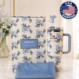 May include: A white Stanley tumbler with a blue bow and floral pattern, a matching box, and a blue cloth. The tumbler has a handle and a straw with a bow-shaped topper. The box has the text "STANLEY LOVESHACKFANCY".