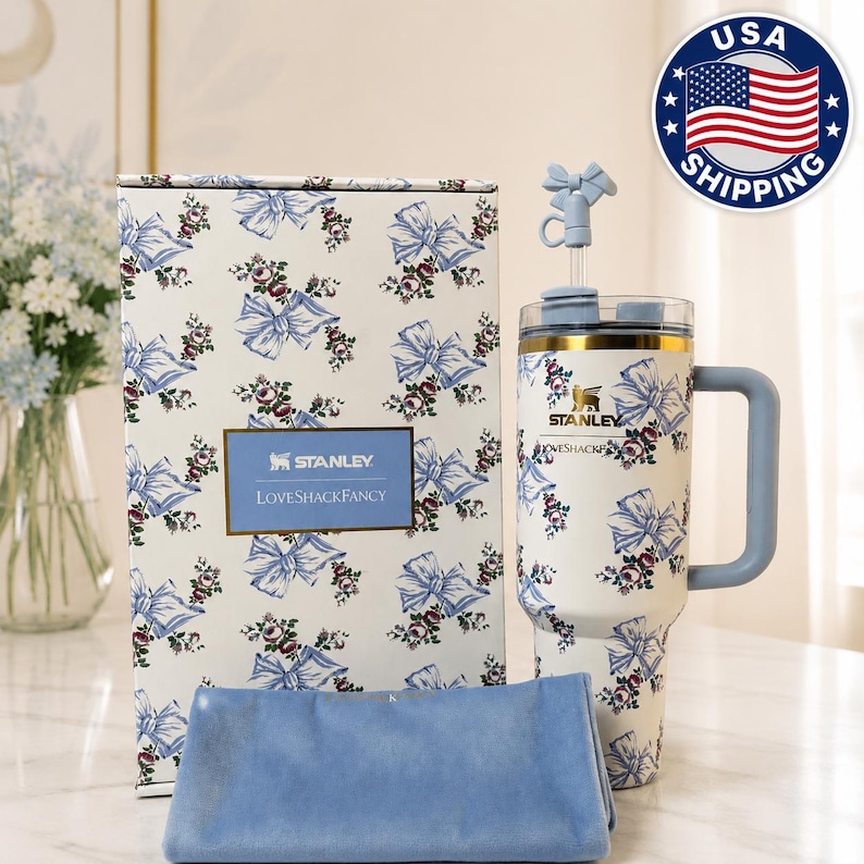 May include: A white Stanley tumbler with a blue bow and floral pattern, a matching box, and a blue cloth. The tumbler has a handle and a straw with a bow-shaped topper. The box has the text "STANLEY LOVESHACKFANCY".