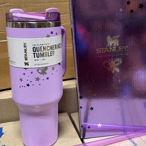May include: A lavender Stanley Quencher H2.0 tumbler with a handle, lid, and straw. The tumbler has a white label with the text "THE FLOWSTATE QUENCHER H2O TUMBLER" and star and butterfly graphics. A matching box is next to the tumbler.