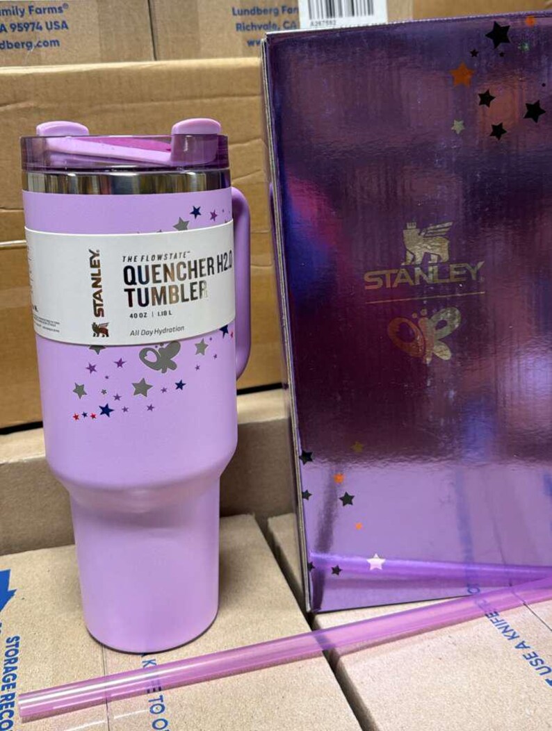 May include: A lavender Stanley Quencher H2.0 tumbler with a handle, lid, and straw. The tumbler has a white label with the text "THE FLOWSTATE QUENCHER H2O TUMBLER" and star and butterfly graphics. A matching box is next to the tumbler.
