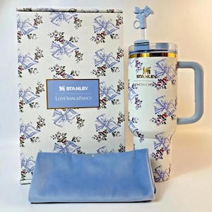 May include: A white Stanley tumbler with a floral and bow pattern, a light blue handle, and a matching straw. The tumbler is paired with a matching box and a light blue cloth. The box has the text "Stanley Loveshackfancy".