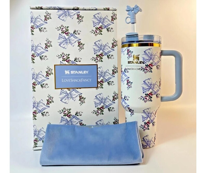 May include: A white Stanley tumbler with a floral and bow pattern, a light blue handle, and a matching straw. The tumbler is paired with a matching box and a light blue cloth. The box has the text "Stanley Loveshackfancy".