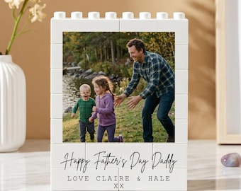 May include: White block photo frame with a family photo. The frame reads "Happy Father's Day Daddy" with names below. A white vase with flowers and a framed abstract art piece are in the background.