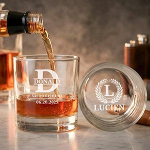 以下が含まれることがあります： 琥珀色の液体が注がれている透明なウイスキーグラス。グラスには「DONALD」という名前、「Groomsmen」という言葉、そして「06.20.2025」という日付が刻まれています。2つ目のガラス蓋には「LUCIEN」という名前が刻まれています。