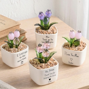 Pode incluir: Quatro pequenos vasos quadrados brancos com esculturas de flores de vidro. Cada vaso tem uma mensagem diferente, como "I Love You Mom" e "Thank You for Helping Me Grow". As flores são rosa e roxas.