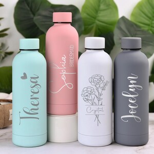 May include: Four insulated water bottles in pastel colors. The bottles are light blue, pink, white, and gray. Each bottle has a name or word engraved on it. The pink bottle says "Sophia Bridesmaid". The white bottle has a floral design.