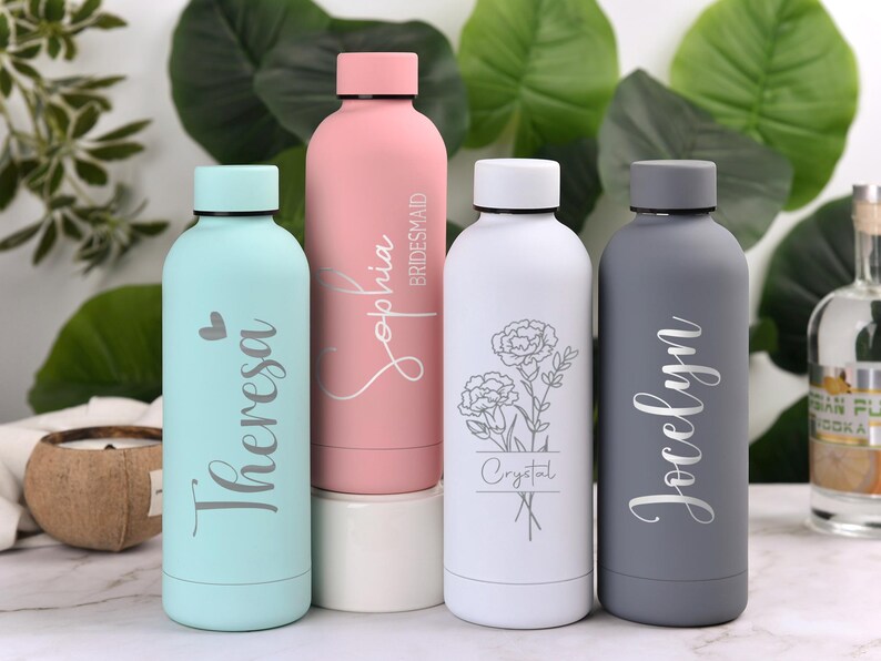 May include: Four insulated water bottles in pastel colors. The bottles are light blue, pink, white, and gray. Each bottle has a name or word engraved on it. The pink bottle says "Sophia Bridesmaid". The white bottle has a floral design.