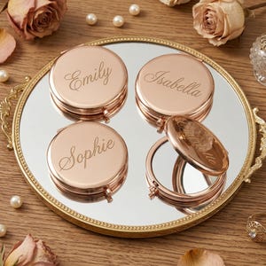 May include: A rose gold compact mirror with the names "Emily", "Isabella", and "Sophie" engraved on the top. The mirror is on a gold-rimmed mirror tray, surrounded by pearls and dried roses. One mirror is open.