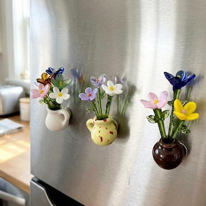 May include: Three glass flower magnets in small vases are attached to a stainless steel refrigerator. The flowers are various colors, including pink, yellow, blue, and white. The vases are white, yellow, and brown.