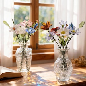 May include: Two glass vases filled with colourful artificial flowers on a wooden table. The flowers include white, pink, blue, and orange blooms. An open book and a window with sunlight are in the background.