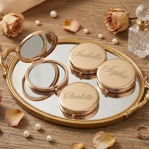 以下が含まれることがあります： 金色のバニティトレイには、コンパクトミラーと、イザベラ、ソフィー、シャーロットの名前が刻まれた3つのパーソナライズされたコンパクトが置かれています。ドライローズ、パール、香水瓶がエレガントなディスプレイを完成させています。