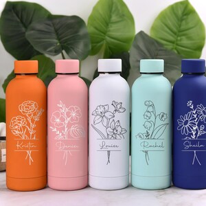 May include: Five colorful insulated water bottles. Each bottle is a different color: orange, pink, white, light blue, and dark blue. Each bottle has a floral design and a name etched on it. The bottles are displayed in front of large green leaves.