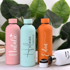 May include: A collection of personalized water bottles in various colors, including pink, light blue, and orange. Each bottle features a name in elegant script. The bottles are cylindrical with a screw-on cap, perfect for hydration on the go.
