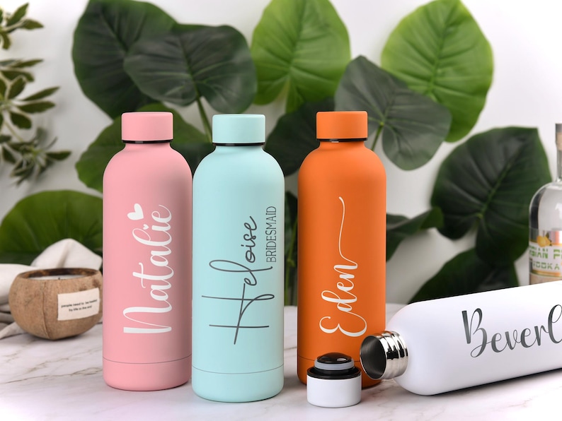 May include: A collection of personalized water bottles in various colors, including pink, light blue, and orange. Each bottle features a name in elegant script. The bottles are cylindrical with a screw-on cap, perfect for hydration on the go.