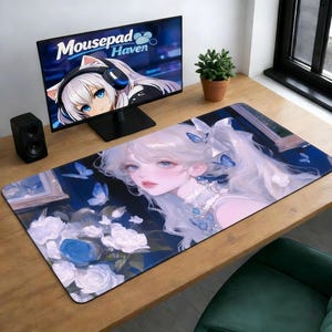 以下が含まれることがあります： 長い白髪、青い目、蝶のアクセントを持つ女性のデジタルイラストが描かれた長方形のマウスパッド。マウスパッドには、白と青のバラの花束もあります。コンピューターモニターには「Mousepad Haven」というテキストが表示されています。