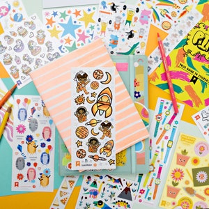 Puede incluir: Una colección de hojas de pegatinas coloridas con varios temas, que incluyen el espacio, los animales y los relojes. Las pegatinas están dispuestas sobre una superficie de color naranja claro.  "Your Guide to Stickers!" está impreso en una de las hojas.