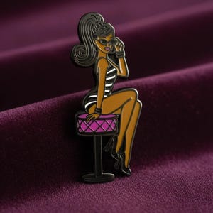 May include: A metal enamel pin featuring a stylised figure with a high ponytail, wearing sunglasses and a black and white striped outfit, sitting on a round pink stool with a diamond-patterned texture and a black base.