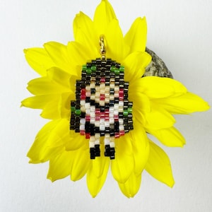 Könnte beinhalten: Ein Perlenanhänger mit einem pixeligen Charakterdesign, mit grünen und schwarzen Haaren, einem roten und weißen Gesicht und einem schwarzen Outfit. Der Anhänger hängt an einem goldfarbenen Verschluss und liegt auf einer leuchtend gelben Blume.