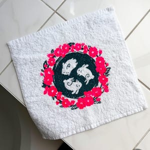 Könnte beinhalten: Ein weißes, quadratisches Handtuch mit einem zentralen kreisförmigen Design. Innerhalb des dunkelgrünen Kreises sind drei weiße Kaninchen kreisförmig angeordnet, umgeben von einem Kranz aus leuchtend pinken Blüten und dunkelgrünen Blättern.
