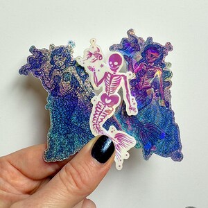 Könnte beinhalten: Drei gestanzte Aufkleber mit zwei holografischen Meerjungfrauen in Blau- und Violett-Glitzeroptik sowie einer zentralen weißen Skelett-Meerjungfrau mit rosa Akzenten, die jeweils detaillierte aquatische Motive zeigen.