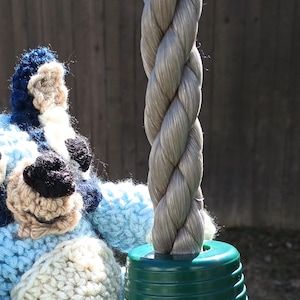 Könnte beinhalten: Ein handgefertigtes Bluey-Plüschtier aus Häkelarbeit in Blau-, Weiß- und Beigetönen. Das Plüschtier sitzt auf einem grünen Schaukelgestell. Das Seil der Schaukel ist grau.
