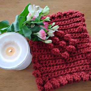Puede incluir: Un cuadrado de crochet rojo con un patrón texturizado y abultado. El cuadrado está sobre una superficie de madera junto a una vela blanca y un ramo de flores rosas y blancas.
