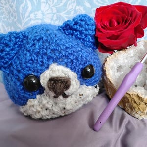 Pode incluir: Um animal de pelúcia azul de crochê com olhos castanhos e um nariz castanho. O animal está sentado em uma superfície roxa ao lado de uma rosa vermelha, um gancho de crochê roxo e um cristal branco.