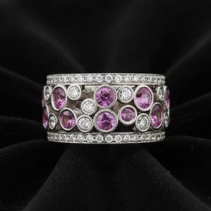 Peut inclure: Une large bague en argent ornée d'un groupe central de saphirs roses ronds et de diamants blancs transparents sertis clos de tailles variées. Les bords supérieur et inférieur sont bordés d'une rangée de petits diamants blancs pavés.