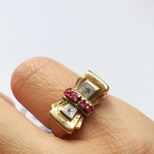 Peut inclure: Une bague dorée avec un motif de nœud papillon. La bague présente une rangée de petites pierres précieuses rouges et un accent de diamant carré. La bague est portée sur un doigt sur fond blanc.