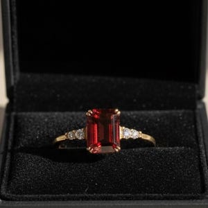 May include: A gold ring with a large, rectangular, red gemstone as the centerpiece. The ring is set with smaller, clear gemstones on either side of the main stone. The ring is displayed in an open black jewelry box.