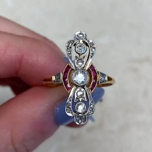 May include: A vintage-style ring with a gold band and an ornate setting. The setting features a central diamond, surrounded by smaller diamonds and red gemstones. The ring's design is symmetrical and elegant, with a classic, timeless appeal.
