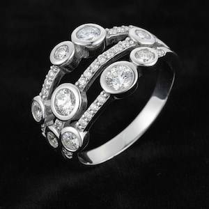 May include: A silver ring with a unique design, featuring multiple rows of sparkling, clear gemstones. The ring has a modern, elegant style, with larger stones set in bezel settings and smaller stones accenting the bands. The ring is set against a black background.