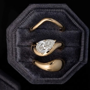 May include: A set of three gold rings with smooth, curved bands. The center ring features a prominent pear-shaped diamond set within a fluid, asymmetrical gold mounting. The top ring has a simple chevron shape, and the bottom ring is a solid gold band.