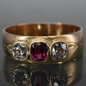 May include: A gold ring with three gemstones. The center stone is a deep red, flanked by two clear, square-cut stones. The ring's band is wide and has subtle decorative etchings. The ring is displayed on a reflective surface.