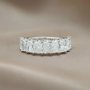 May include: A silver ring featuring a series of radiant-cut, clear gemstones. The gemstones are set closely together, creating a continuous band of sparkle. The ring is displayed on a neutral, slightly textured surface.