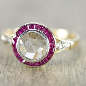 May include: A gold ring with a large, faceted, clear gemstone at the center, surrounded by a halo of small, vibrant red gemstones. The ring has a vintage design with delicate detailing on the band.