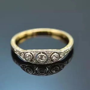 Puede incluir: Un anillo dorado con una banda que presenta tres piedras preciosas centrales, redondas y transparentes. El anillo tiene un diseño decorativo alrededor de las piedras preciosas y a lo largo de la banda. El anillo se muestra sobre una superficie reflectante.