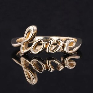 Puede incluir: Un anillo dorado con la palabra "love" en cursiva. El anillo se muestra sobre una superficie negra reflectante, creando un efecto de espejo. Es una pieza de joyería.