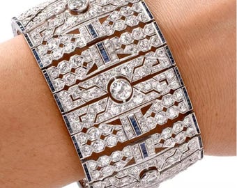 May include: A dazzling, Art Deco-style bracelet. The bracelet features a geometric design with numerous clear, sparkling stones and small blue accents. The bracelet is made of a silver-colored metal and is a statement piece of jewelry.