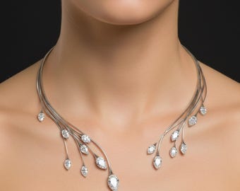Könnte beinhalten: Eine silberne Halskette mit zartem, fließendem Design. Die Halskette hat mehrere Stränge, die sich um den Hals legen und mit tropfenförmigen, klaren Edelsteinen verziert sind. Die Edelsteine variieren in der Größe und sind gleichmäßig verteilt.