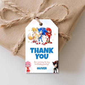 May include: A white gift tag with the words "THANK YOU" in blue, featuring cartoon characters Sonic, Tails, and Knuckles. The tag also includes the text "for coming to my birthday party!" and the name "OLIVER". The tag is tied to a wrapped gift with twine.