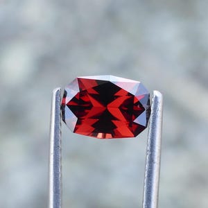 May include: A deep red gemstone with a rectangular cut, held in tweezers. The gemstone is a vibrant red color and has a high level of clarity.