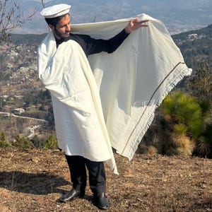 May include: A cream-colored shawl with a fringed edge and a decorative border is held up in the air. The shawl is draped over a person wearing a white cap, black shirt, and black pants. The background features a mountainous landscape.