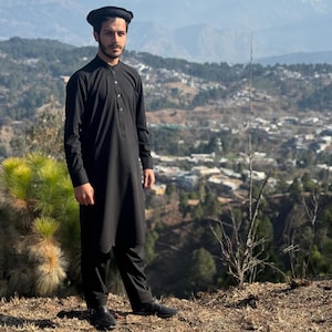 May include: A person wearing a black traditional outfit, including a long-sleeved tunic and matching pants. The outfit is paired with a black cap. The background features a scenic view of mountains and a town.