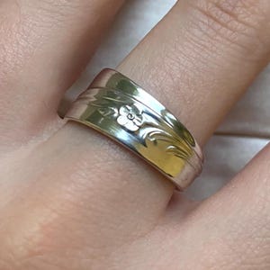 May include: A silver ring with a floral design, worn on a finger. The ring appears to be made from a spoon, with the floral pattern and curved shape of the spoon visible. The ring is a bright, shiny silver colour.