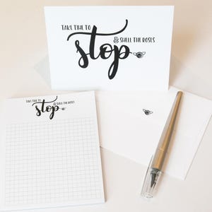 May include: A white card with black text that reads "Take time to stop & smell the roses". The card is accompanied by a white notepad with a grid pattern and the same text in black. A silver pen is also included.